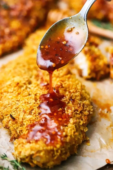 A person using a silver spoon to drizzle honey hot sauce over the top of a crispy corn flake-crusted chicken cutlet.