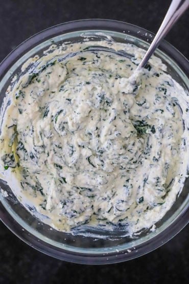 An overhead view of a large glass bowl that is filled with a ricotta and chopped spinach mixture with a large silver spoon inserted off to the side of the bowl.
