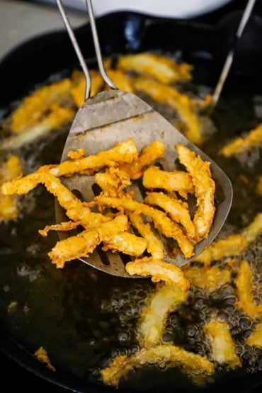 A close-up view of a metal slotted spatula raising crispy fried onion strips from a skillet filled with hot oil.