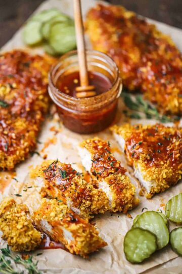 A straight-on view of a cutlet of hot honey chicken that has been cut into strips and drizzled with hot honey sauce and is surrounded by more cutlets and butter pickles.