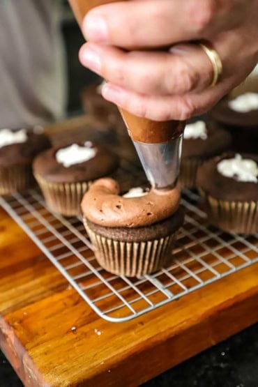 Person using a piping bag fitted with a metal tip to apply chocolate frosting on top of chocolate cupcakes with marshmallow filling all sitting on a baking rack.