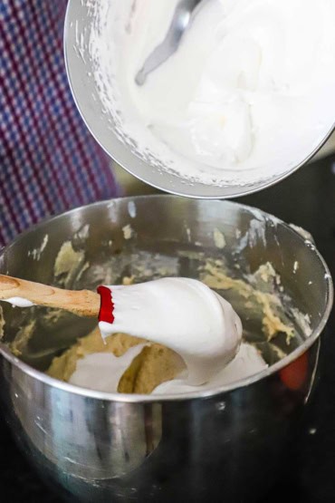 A person using a large red spatula to fold soft peak egg whites into a mixing bowl filled with a white cake batter.
