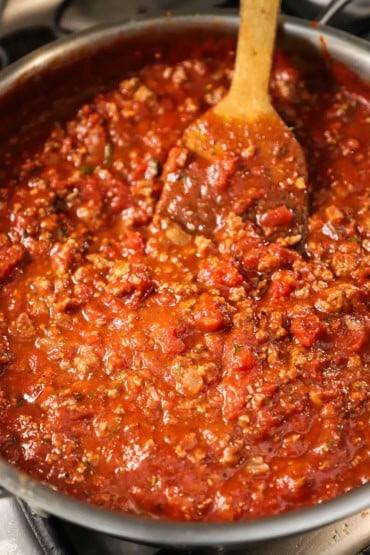 A close-up view of a tomato-based meat sauce in a large silver saucepan with a wooden spatula inserted into the sauce near the back of the pan.