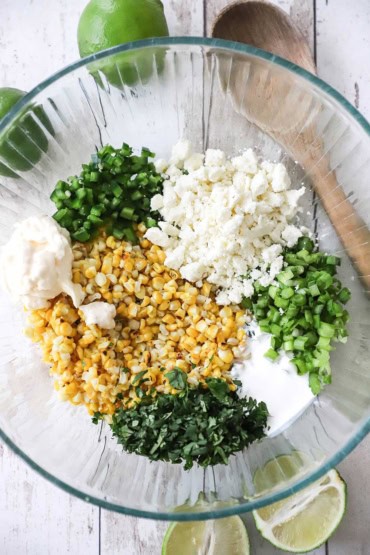 An overhead view of a large glass bowl that is filled with roasted corn, chopped cilantro, jalapeño, scallions, crumbled Cotija cheese, mayonnaise, and Mexican crema.