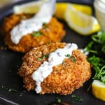 A straight-on view of two New England-style crab cakes that are on a black dinner plate both with a strip of tartar sauce on them and sitting next to lemon wedges.