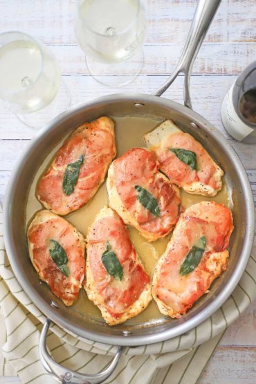 An overhead view of six fully cooked chicken saltimbocca cutlets in a skillet that are topped with crispy prosciutto and a sage leaf all resting in a white wine butter sauce.