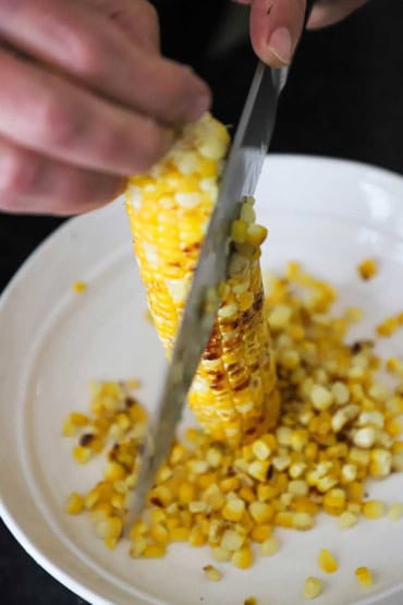 A close-up view of a person using a large knife to cut the lightly charred kernels from a cooked ear of corn in a shallow white bowl.