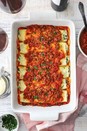 An overhead view looking straight down on a white 9 by 13 baking dish that is filled with spinach and ricotta cannelloni topped with a tomato-based meat sauce.