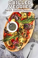 An overhead view of a wooden oval platter filled with grilled vegetables that have been drizzled with Italian dressing in a white bowl with a spoon on it.