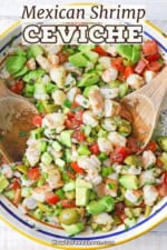 An overhead view of a large colorful bowl filled with Mexican shrimp ceviche with two wooden spoons inserted into the sides of the dish.