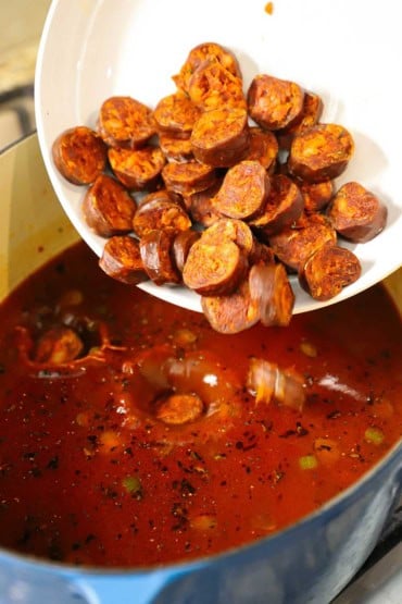 A person dumping sliced Spanish chorizo from a shallow white bowl into a large oval Dutch oven filled with a tomato-based broth.