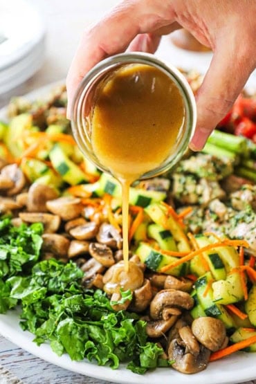 A person pouring a red wine vinaigrette from a small glass jar over a platter filled with rows of grilled mushrooms, cucumber salad, and grilled chicken pieces.