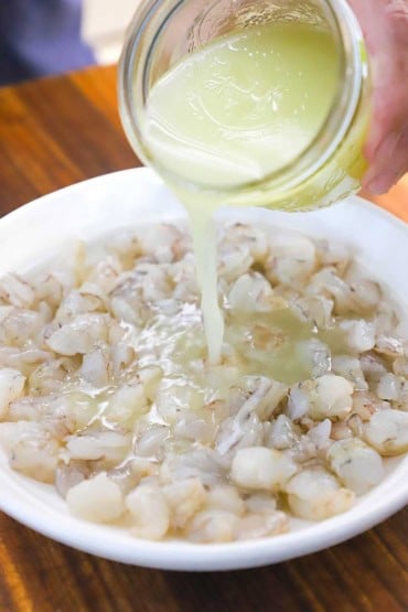 A person pouring freshly squeezed lime juice from a glass jar into a shallow white bowl that is filled with raw shrimp that has been cleaned and cut into bite-sized pieces.