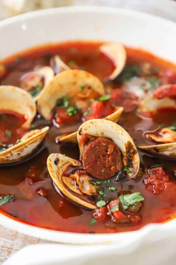 A close-up view of a white bowl filled with a serving of Portuguese-style clam chowder with pieces of cooked chorizo, tomato chunks, and opened littleneck clams visible.