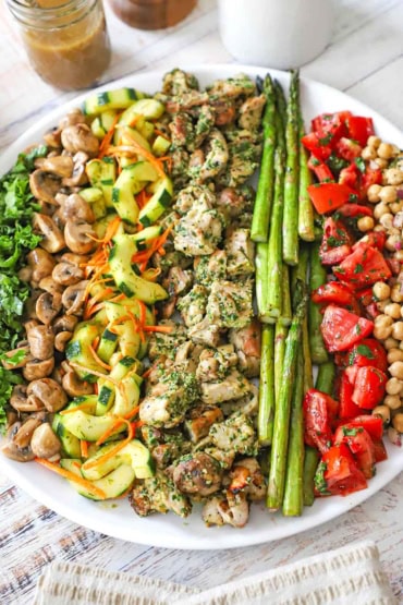 A close-up view of a grilled chicken salad with vegetables on a large white platter with a jar of vinaigrette sitting nearby.