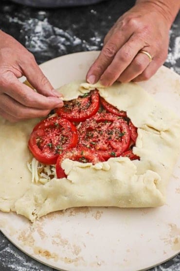 A person folding over homemade pie dough over a pile of caramelized onions, shredded Gruyere cheese, and sliced tomatoes.
