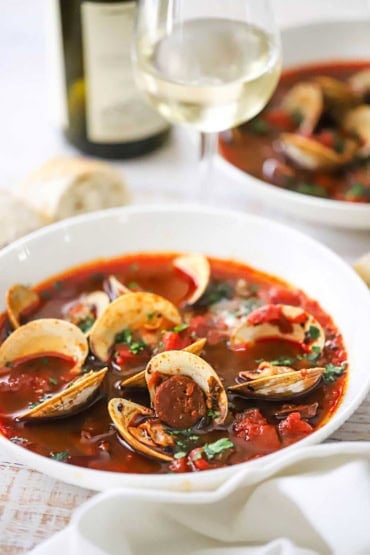 A straight-on view of a two white bowls filled with Portuguese-style clam chowder both with a glass of white wine nearby.
