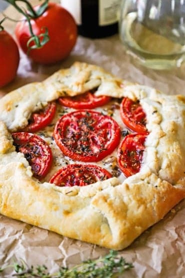 A straight-on view of a baked tomato galette with gruyere cheese and caramelized onions sitting next to a sprig of thyme and two tomatoes on a vine.