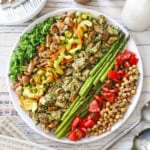 An overhead view of a grilled chicken salad salad that consists of rows of grilled pesto chicken, asparagus, mushrooms, and salads of cucumbers, tomatoes, and chickpeas.
