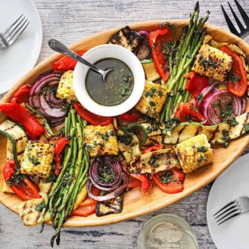 An overhead view of a wooden oval platter filled with grilled vegetables that have been drizzled with Italian dressing in a white bowl with a spoon on it.