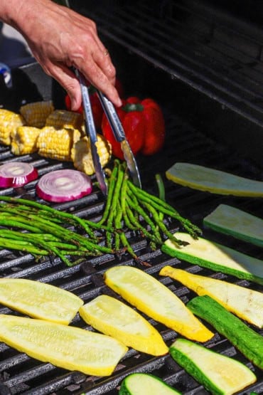 A person using a pair of metal tongs to place thin asparagus on the grates of a gas grill along side strips of zucchini, squash, red onion, as well as corn on the cob and whole red bell peppers.