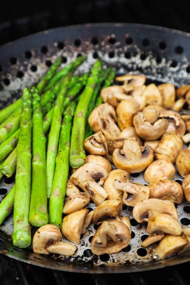 A close-up view of a grill pan on a gas grill and is holding a pile of asparagus and quartered pieces of white button mushrooms.