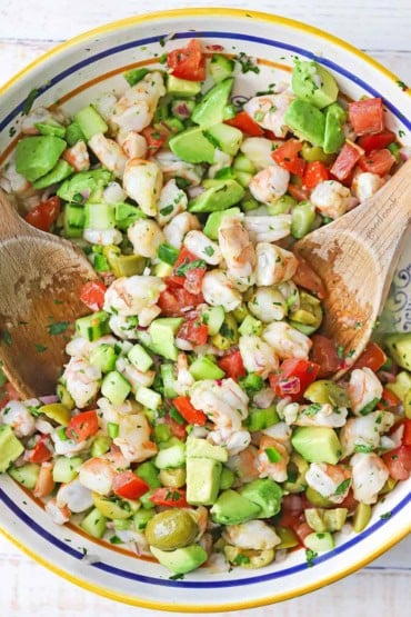 An overhead view of a large colorful bowl filled with Mexican shrimp ceviche with two wooden spoons inserted into the sides of the dish.