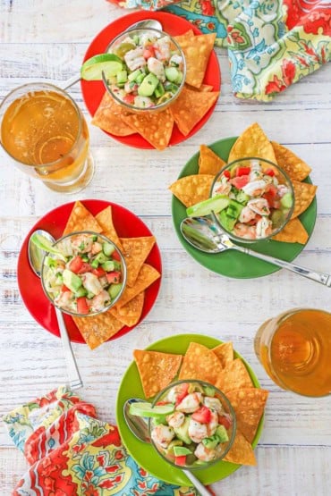 An overhead view of four servings of shrimp ceviche in glass bowls resting on colorful cocktail plates with corn tortilla chips and two glasses of beer nearby.