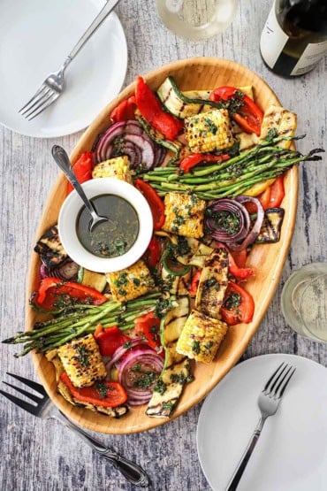 An overhead view of a wooden oval platter filled with grilled vegetables that have been drizzled with Italian dressing in a white bowl with a spoon on it.