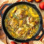 An overhead view of large black cast-iron skillet that is filled with braised chicken and peaches with a wooden spoon and fresh peaches nearby.