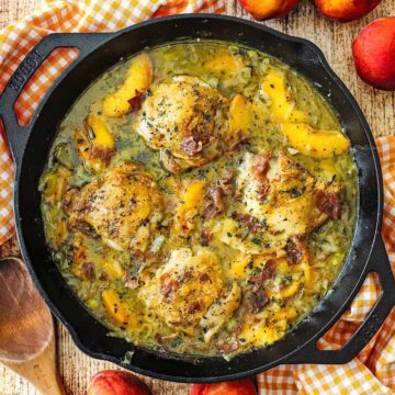 An overhead view of large black cast-iron skillet that is filled with braised chicken and peaches with a wooden spoon and fresh peaches nearby.
