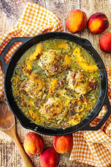 An overhead view of large black cast-iron skillet that is filled with braised chicken and peaches with a wooden spoon and fresh peaches nearby.