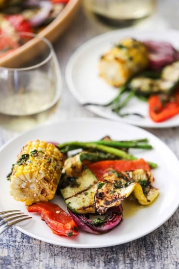 A straight-on view of two cocktail plates that are filled with grilled piece of corn on the cob, slices of squash, red onion, and asparagus.