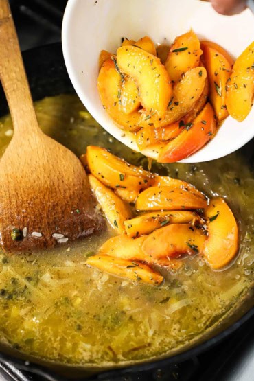 A person transferring a bowl filled with marinated peach slices into a skillet filled with a simmering broth and sautéed fennel strips.