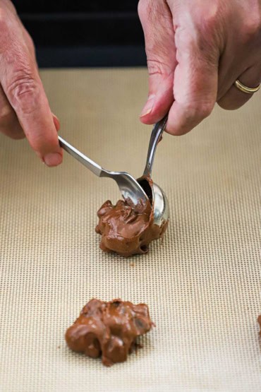 A person using a small spoon to scrape chocolate cookie dough out of a tablespoon onto a silicone baking matt.