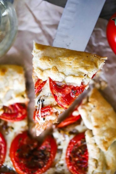 A sliced of a tomato galette being raised up on a large stainless steel knife.