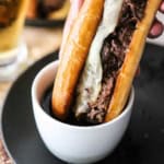 A close-up view of a slow cooker French dip sandwich being plunged into a small bowl of au jus sauce on a black plate.