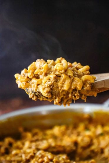 A large wooden spoon being raised from a skillet that is filled with homemade cheesy hamburger helper with steam rising into the air above it.