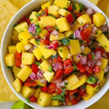 A close-up view of mango salsa that is in a white bowl with tortilla chips and lime wedges surrounding it.