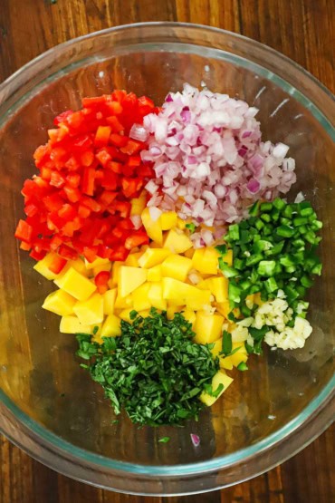 An overhead view of a large glass bowl that is holding small piles of chopped mango, red bell pepper, red onion, jalapeño, garlic, and cilantro.