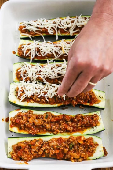 A person sprinkling shredded mozzarella cheese over the tops of halved zucchinis that have been filled with a sausage and tomato sauce.
