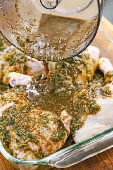 A person pouring Jamaican jerk marinade over whole chicken legs in a large glass baking dish.