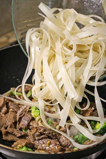 A person transferring softened wide rice noodles into a wok that is filled with stir-fried steak and broccoli.