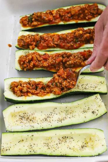 A person spooning an Italian sausage and tomato sauce onto zucchini boats that have been cut in half, lengthwise, and the seeds carved out.