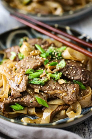 A close-up view of a colorful dinner plate filled with a serving of beef and broccoli chow fun with two wooden chop stick resting on the side of the plate.