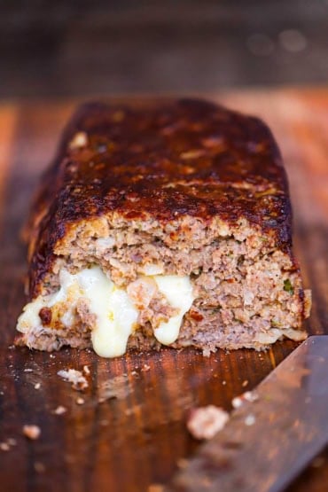A straight-on view of a smoked meatloaf that has the end cut away and melty cheese on the inside of the loaf is visible and oozing down the front of the sliced loaf.