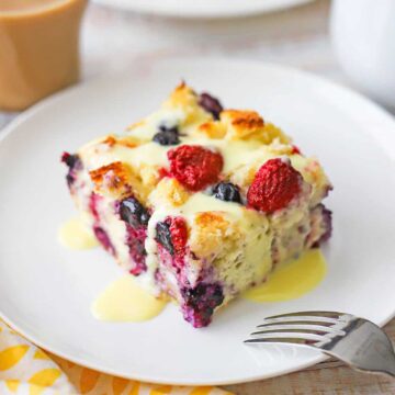 A straight-on view of a slice of mixed berry bread pudding with orange sauce sitting on a white dessert plate with a glass mug of creamed coffee in the background.