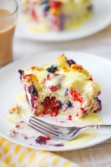 A straight-on view of a square piece of mixed berry bread pudding and orange sauce with a bite missing from it and a fork resting on the plate.