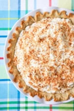 An overhead view of a coconut cream pie that is untouched in a white pie dish and sitting on a colorful place setting.