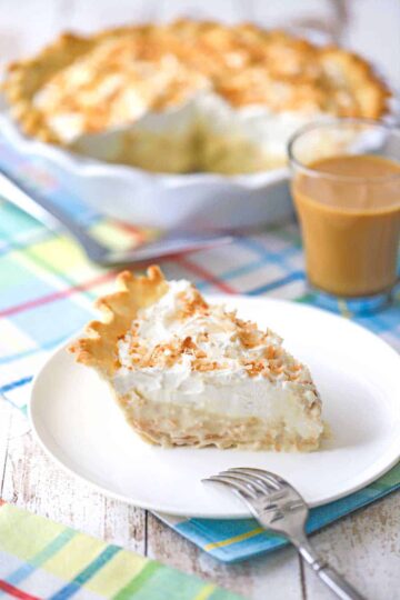 A close-up view of a slice of coconut cream pie sitting on a white dessert plate with a glass mug off coffee in the background.